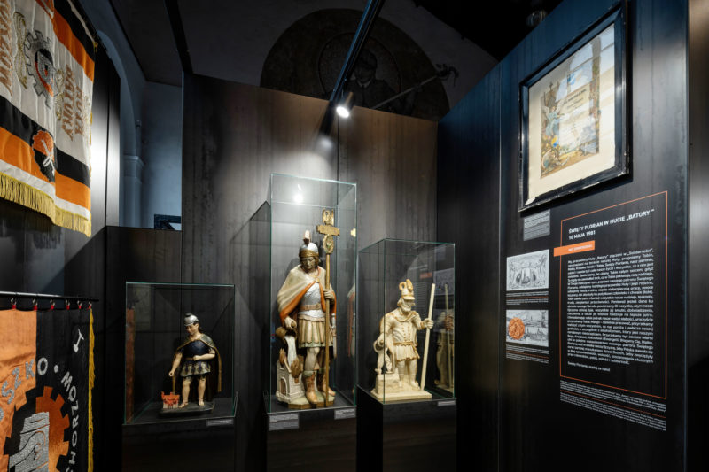Trzy figurki św. Florianów o różnych wielkościach. Trzymają dzban z wodą polewając budynek, dwóch z nich trzyma laskę. Po prawej opis wystawy, z lewej dwa sztandary.