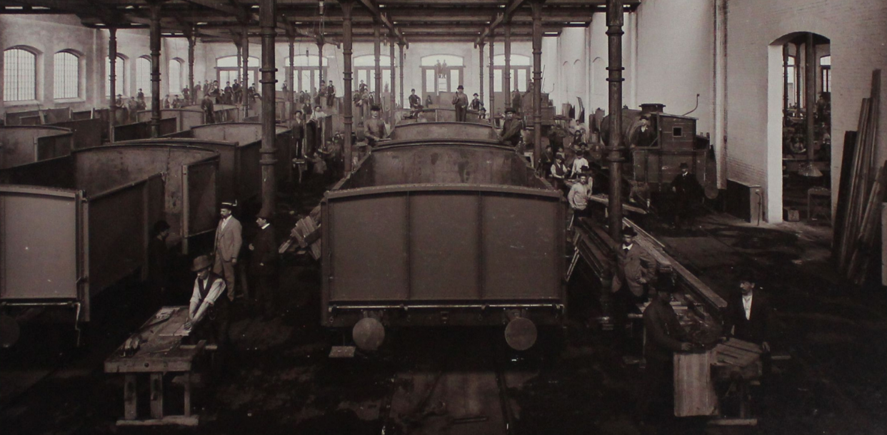Wnętrze hali produkcyjnej z wagonami, obok pracownicy oraz materiały budowlane. Część pracowników stoi na wagonach. Duża sala z wielkimi oknami