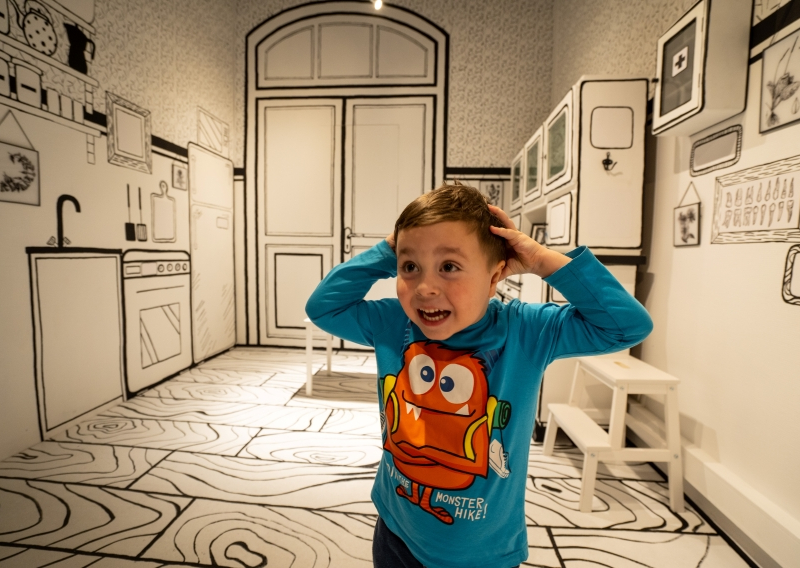Dziecko trzymające się za głowę w kolorowej koszulce. Znajduje się w pomieszczeniu inscenizującym pokój. Na ścianie po lewej narysowana czarną linia na białym tle kuchnia oraz urządzenia jakie się w niej znajdują. Po prawej szafka z apteczką i obrazy. Z tyłu drzwi wejściowe