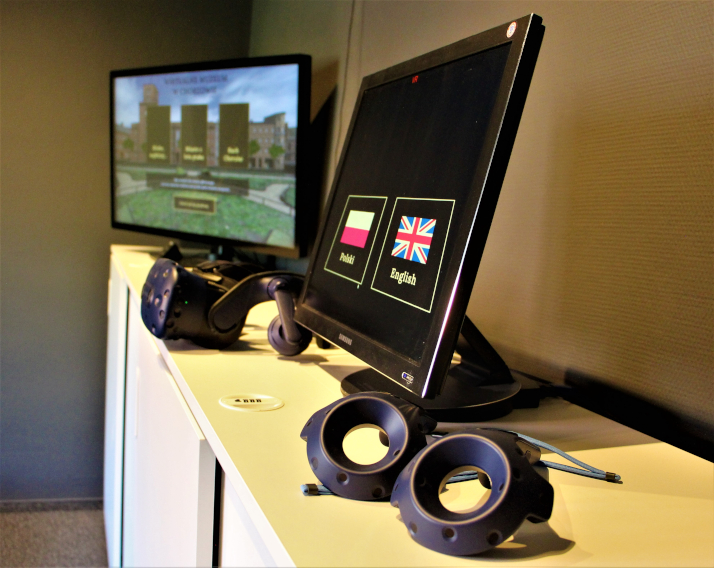Stolik z dwoma monitorami i goglami VR. Na pierwszym ekranie widoczny jest wybór języka z flagą, na drugim budynek.
